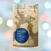 A package of Glyde Maxi LARGE Size Vegan Condoms, from the brand Glyde, showcases its vegan origins and eco-friendly production with packaging crafted from natural ingredients. The design is accentuated by blurred, colorful bokeh lights in the background, adding a vibrant touch.